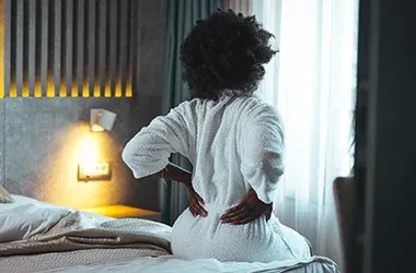 Person sitting on the edge of a bed holding their lower back with both hands